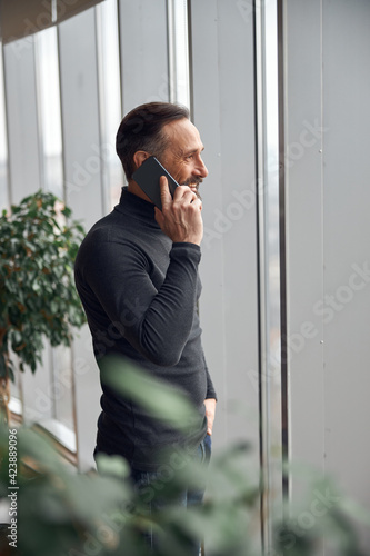 Happy smart adult caucasian businessman is standing near big windows in office space and holding smartpone