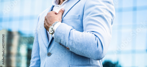 Watch in a man. Businessman points to his watch on the background of the town. Man holds his watch. Portrait successful businessman in a business suit, using the watch on a city background