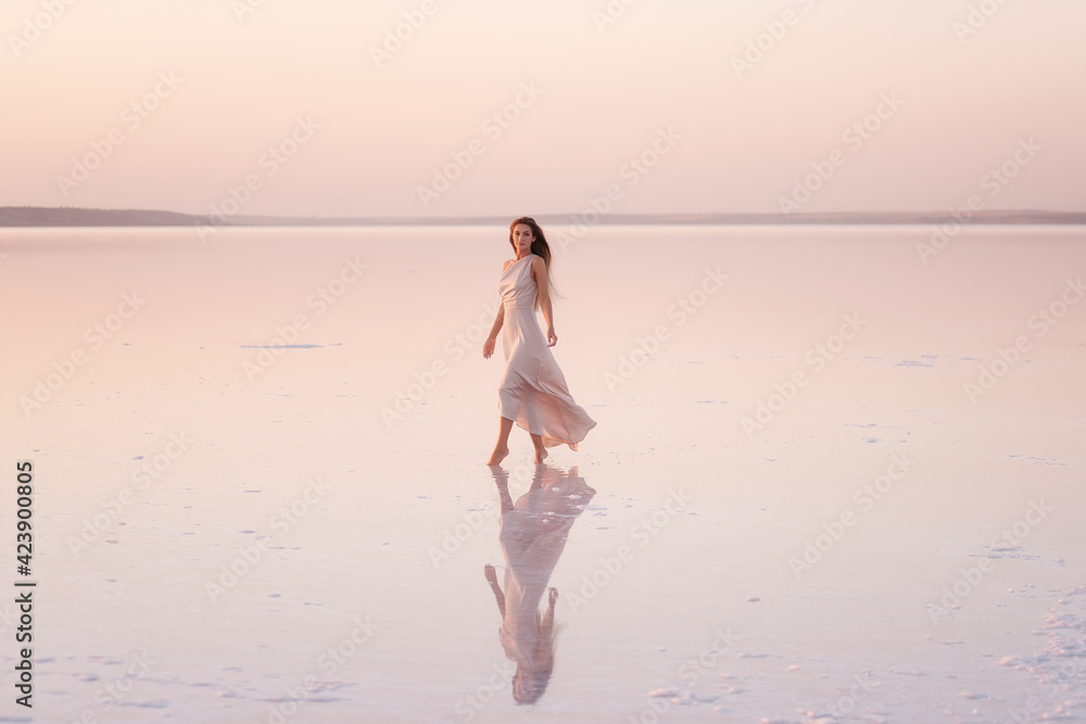 Young blonde woman in an evening airy pastel pink, powdery dress stands barefoot on white crystallized salt. Girl with natural make-up, hair is developing. Salt mining trip, walking on water at sunset Young blonde woman in an evening airy pastel pink, powdery dress stands barefoot on white crystallized salt. Girl with natural make-up, hair is developing. Salt mining trip, walking on water at sunset