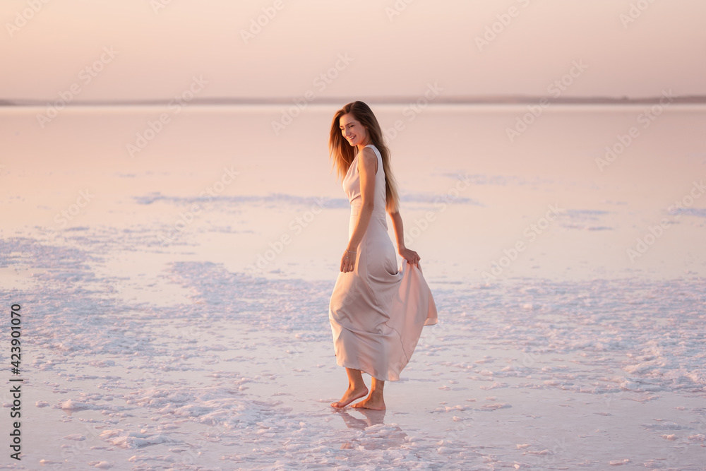 Young blonde woman in an evening airy pastel pink, powdery dress stands barefoot on white crystallized salt. Girl with natural make-up, hair is developing. Salt mining trip, walking on water at sunset Young blonde woman in an evening airy pastel pink, powdery dress stands barefoot on white crystallized salt. Girl with natural make-up, hair is developing. Salt mining trip, walking on water at sunset