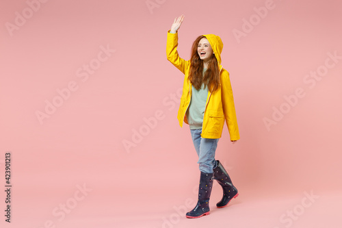 Full length redhead fun young woman in yellow waterproof hood raincoat outerwear walk go waving hand greet isolated on pastel pink background studio Outdoors lifestyle wet fall weather season concept.