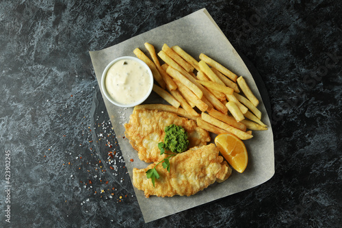 Canvas Print Baking paper with fried fish and chips on black smokey table
