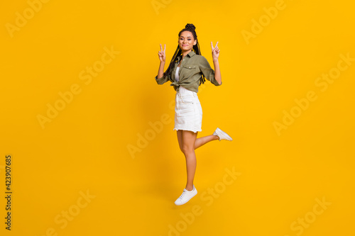 Photo portrait full body view of dark skin woman showing v-signs jumping up isolated on vivid yellow colored background