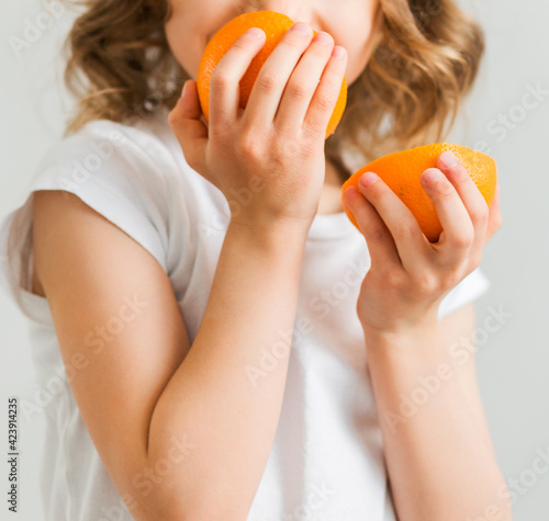 A little girl in a white T-shirt holds two orange slices in her hands and sniffs them