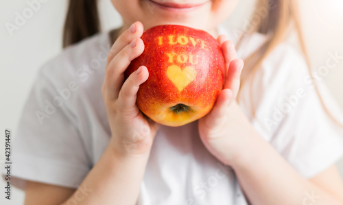 A little girl in a white T-shirt holds an apple in her hands with the inscription I love you