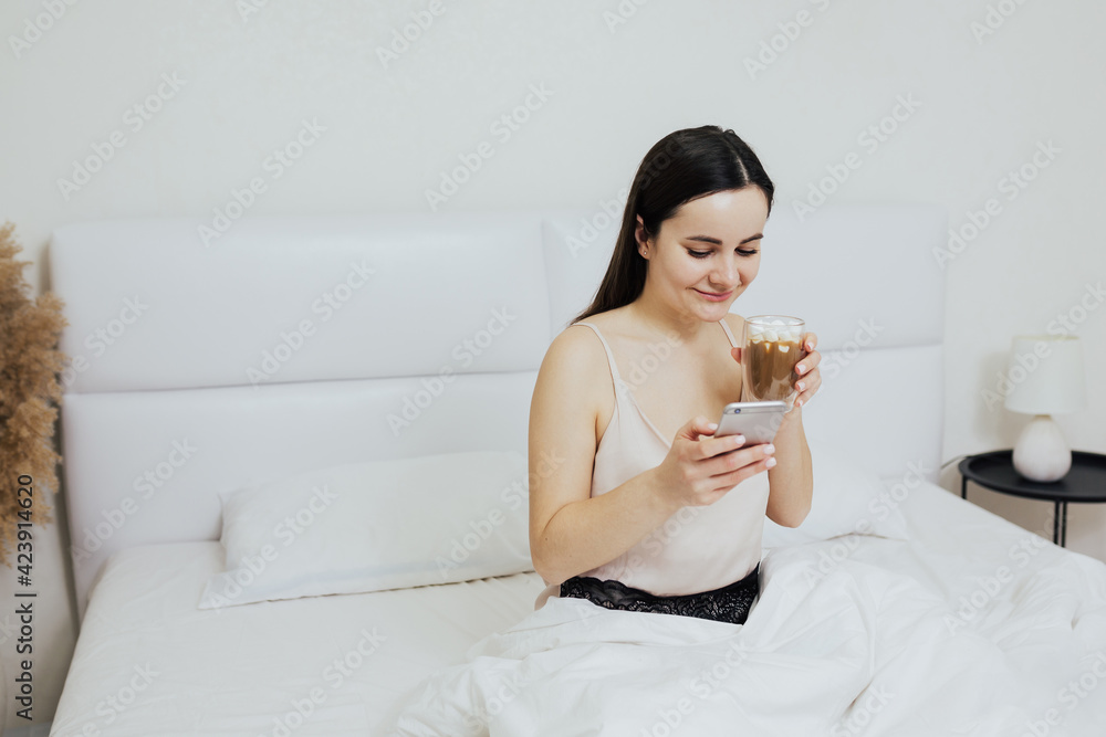 Happy young beautiful woman in pajamas drinks cappuccino while using smartphone, sitting on the bed. 