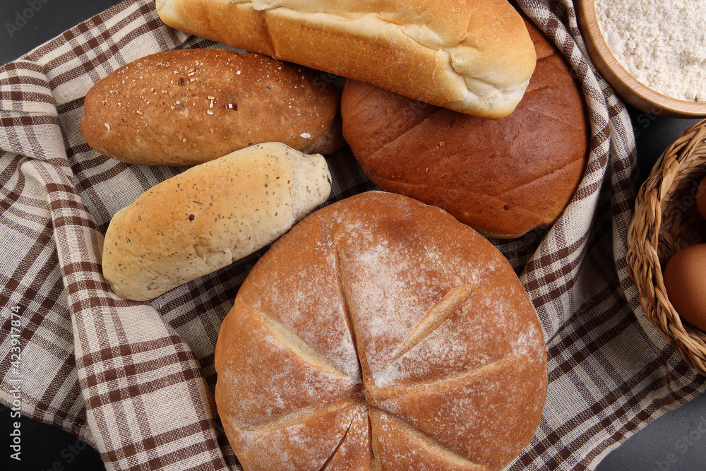 Freshly baked bread loaf bun roll round long mix verity wrapped in checkered kitchen fabric napkin towel wheat flower oil water salt eggs rolling pin slate stone over black background