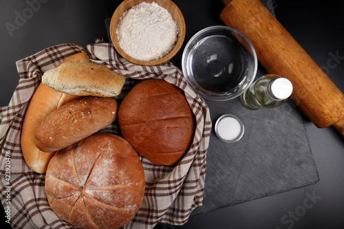 Freshly baked bread loaf bun roll round long mix verity wrapped in checkered kitchen fabric napkin towel wheat flower oil water salt eggs rolling pin slate stone over black background photo