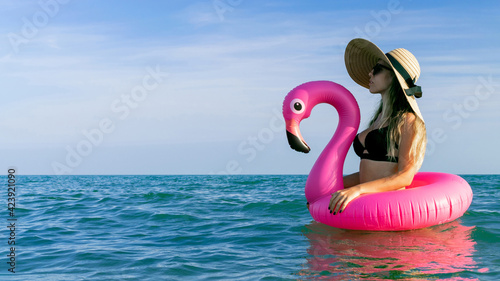 Summer hat beach. Happy young sexy girl in bikini swimsuit, sunglasses and straw hat with pink inflatable flamingo in blue sea water on ocean background. Luxury lifestyle travel. © Maksym