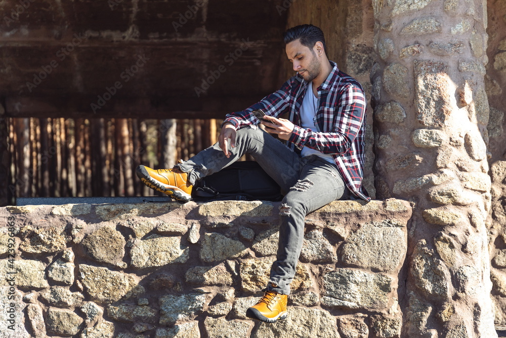Fototapeta premium Attractive man sitting on stone wall looking at mobile phone.