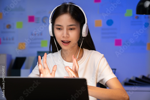 Cheerful beautiful Asian woman in a white t-shirt with wearing headphones and look to laptop at video calling meeting and study online on the Internet.