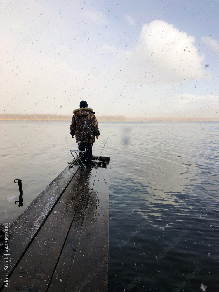 A woman in warm clothes on the shore of the lake fishing with a fishing rod. It is snowing in large flakes. The fisherman stands on an old wooden pier. View from the back.