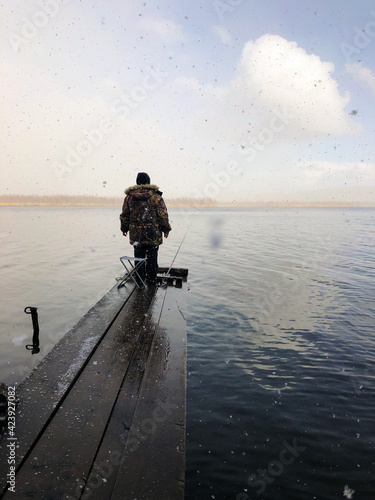 A woman in warm clothes on the shore of the lake fishing with a fishing rod. It is snowing in large flakes. The fisherman stands on an old wooden pier. View from the back.