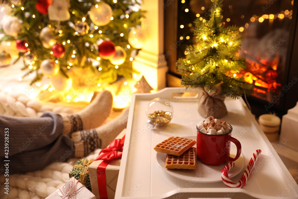 Woman sitting near fireplace at home, focus on tray with cocoa and snacks. Cozy winter holidays atmosphere Woman sitting near fireplace at home, focus on tray with cocoa and snacks. Cozy winter holidays atmosphere
