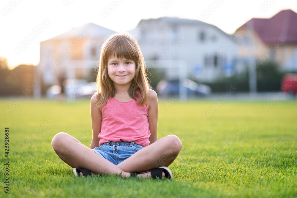 Young pretty child girl sitting on fresh green grass lawn on warm summer day outdoors.