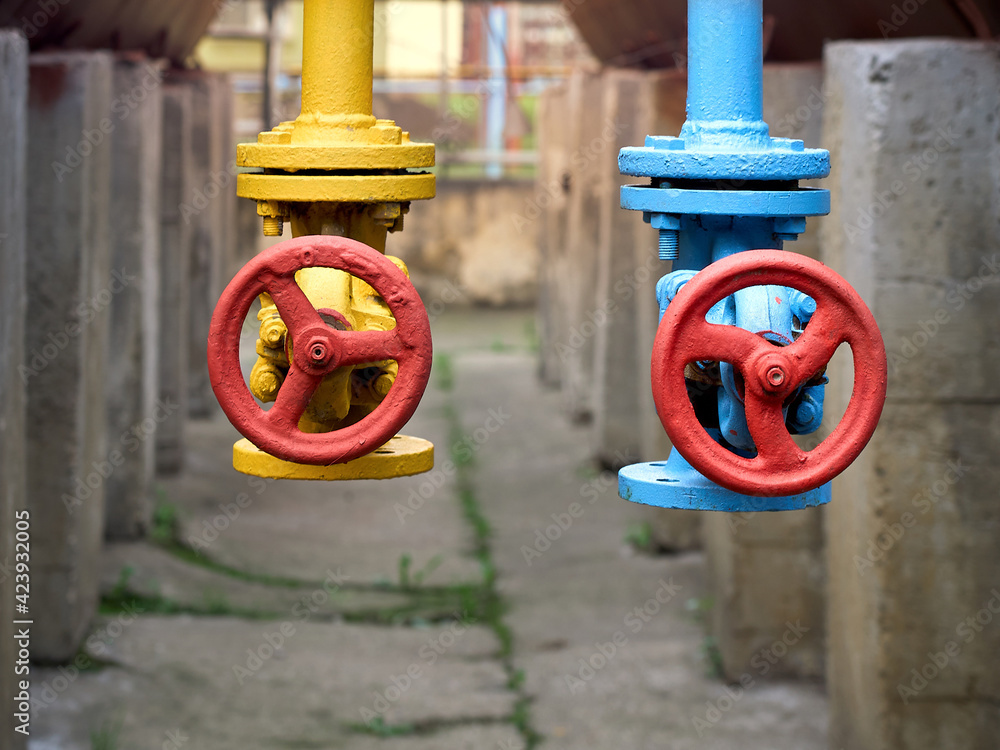 Two drain valves blue and yellow with red hand wheels over concrete elements and brown tanks background. Steampunk retro 1980s design industrial petrochemical chemical plant with sunflare background.
