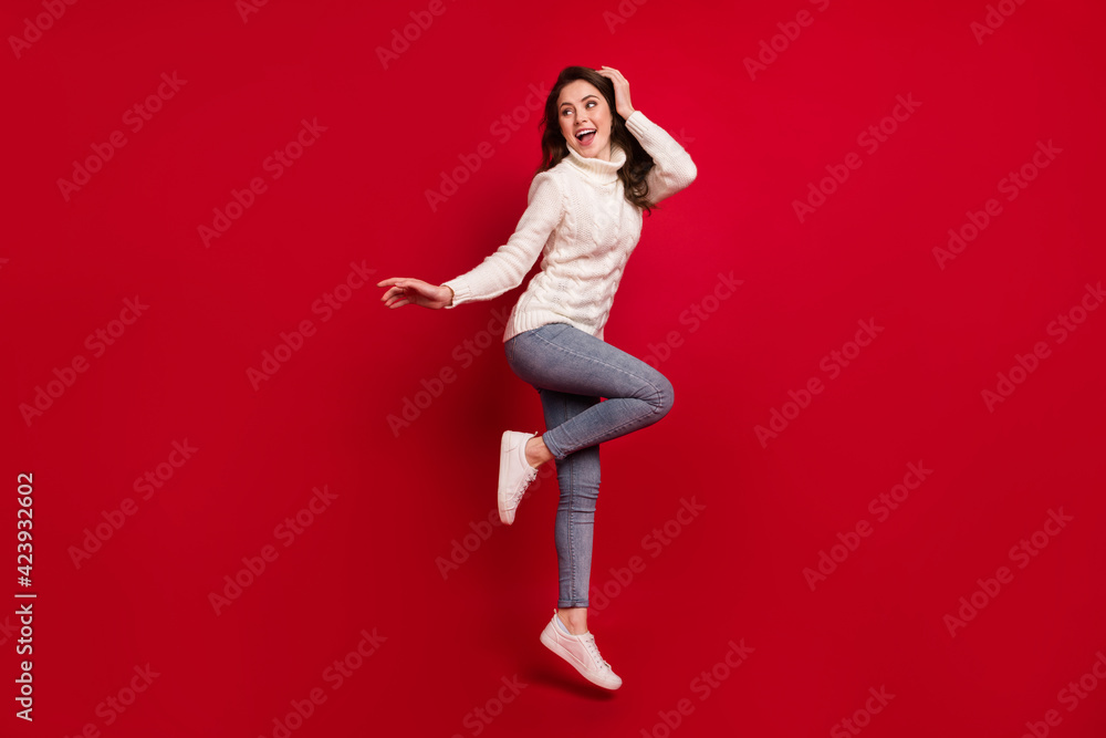 Full length photo of pretty shiny young woman dressed white sweater jumping high arm head isolated red color background Full length photo of pretty shiny young woman dressed white sweater jumping high arm head isolated red color background