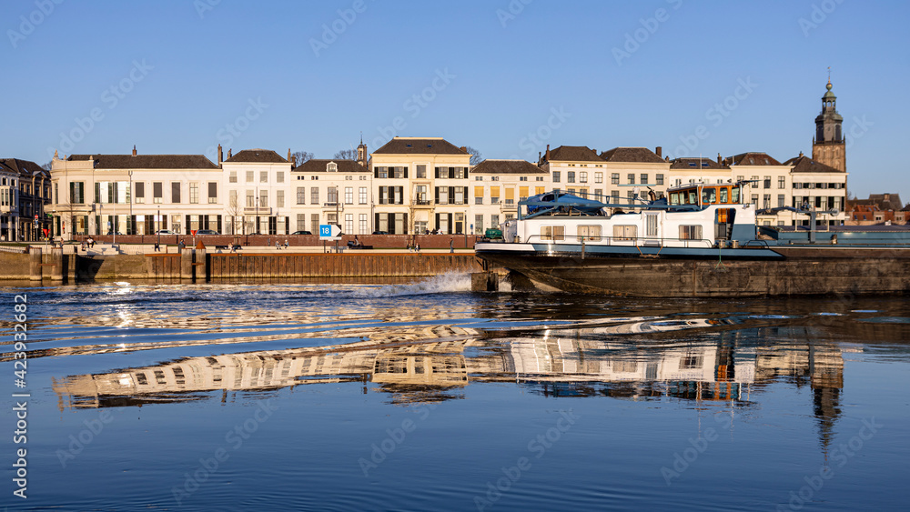 Cargo ship speeding past the IJsselkade boulevard of Hanseatic city of Zutphen, The Netherlands, traversing through the reflection of the cityscape in the water. Countenance panorama of Dutch city.