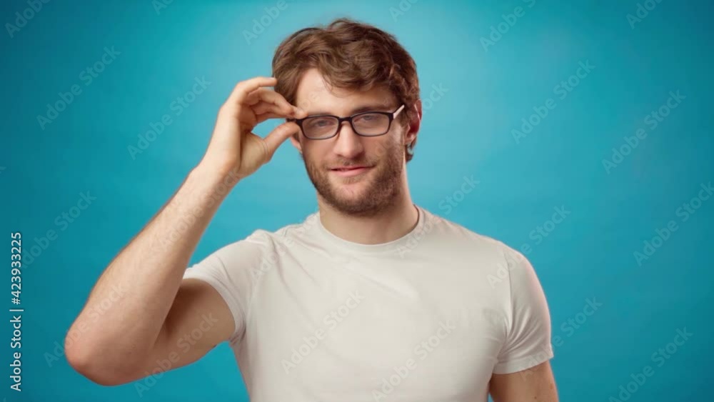 Young man taking off his glasses slowly against blue background Young man taking off his glasses slowly against blue background