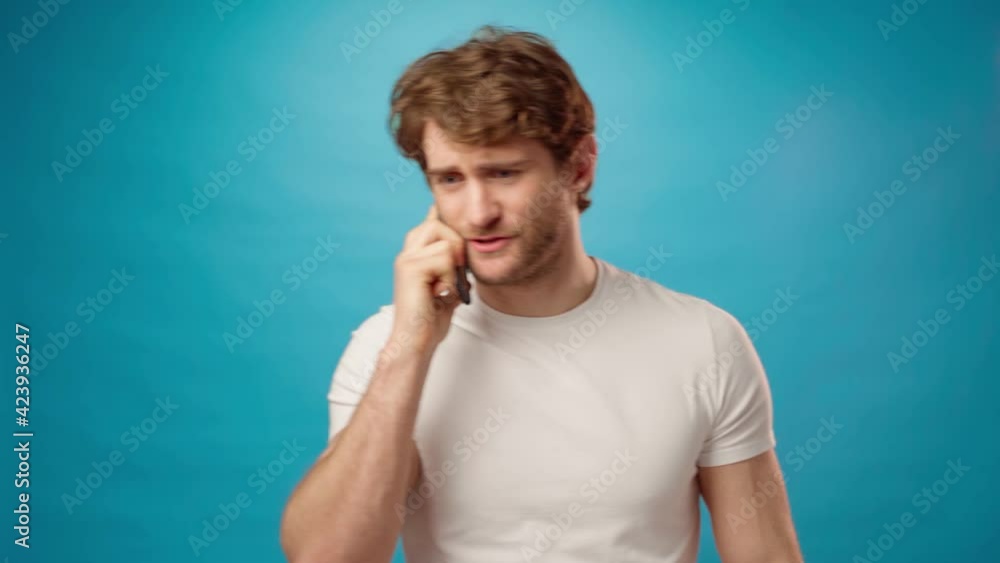 Angry irritated young man in white shirt talking on the phone against blue background Angry irritated young man in white shirt talking on the phone against blue background
