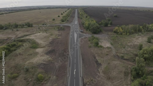 The copter flies over the road, to the left and right of it is a field and a forest, a car is driving along the road