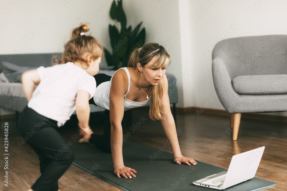 Obraz premium A young mom is doing sports at home and watching a video fitness lesson on a laptop, and her little baby is sitting on a chair. Home training, fitness, sports