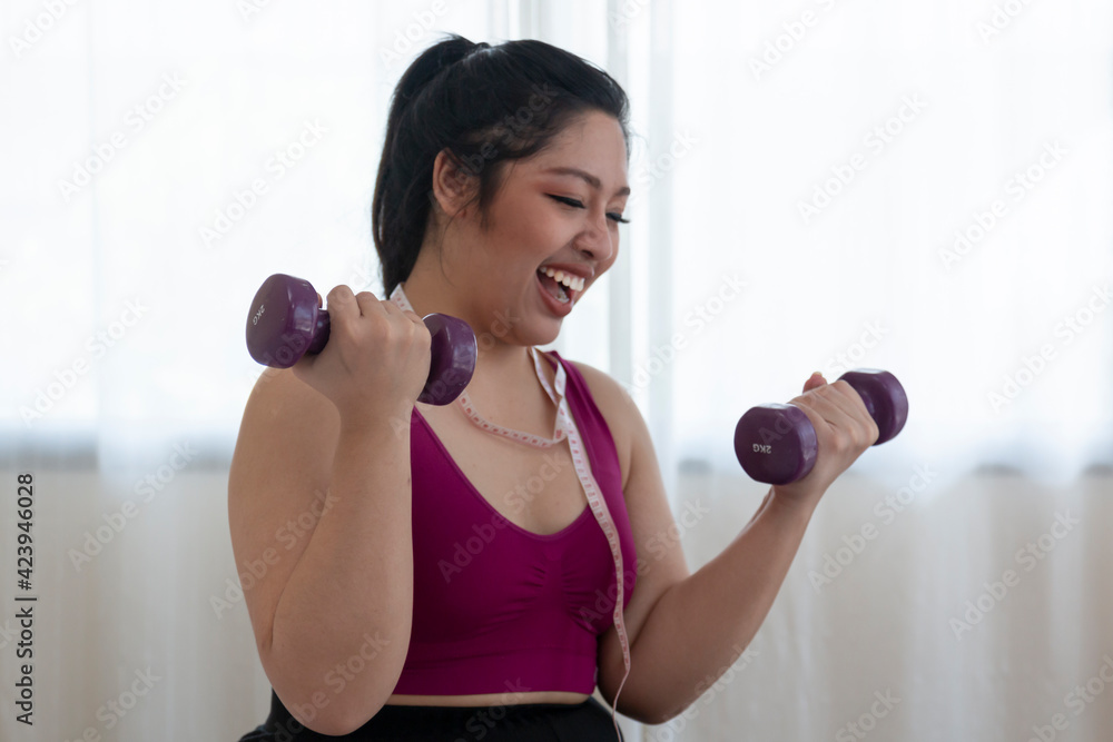 Selected focus at dumbbell. Overweight woman doing exercise with dumbbells at home. Overweight fat woman with measure tape on neck holding dumbbell in hands while sitting at home. Selected focus at dumbbell. Overweight woman doing exercise with dumbbells at home. Overweight fat woman with measure tape on neck holding dumbbell in hands while sitting at home.