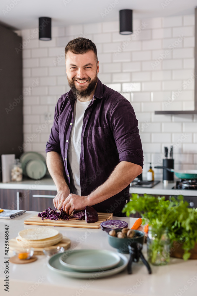 Portrait of a Handsome Man slicing Red Cabbage on a Cutting Board and preparing Healthy Fresh Food with Organic Vegetables at Home in Cozy Kitchen, Copy Space Portrait of a Handsome Man slicing Red Cabbage on a Cutting Board and preparing Healthy Fresh Food with Organic Vegetables at Home in Cozy Kitchen, Copy Space