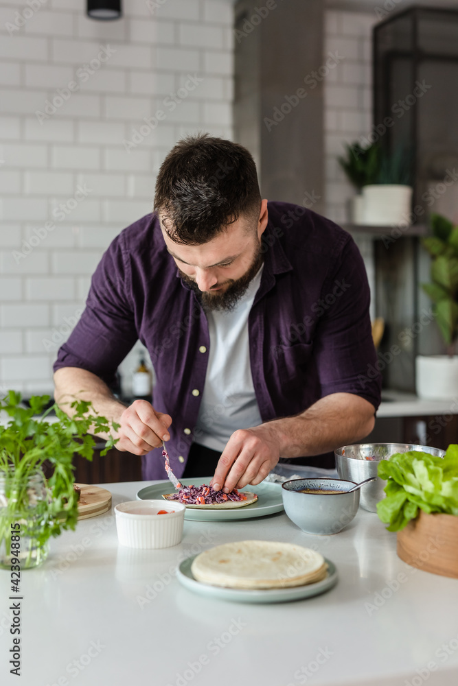 Man in Kitchen preparing Mexican Food with Healthy Ingredients and Vegetables adding Salad on Tortilla at Home on Daylight, Portrait, Copy Space Man in Kitchen preparing Mexican Food with Healthy Ingredients and Vegetables adding Salad on Tortilla at Home on Daylight, Portrait, Copy Space