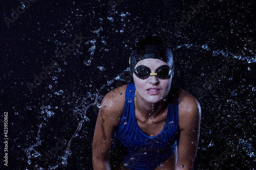Sportive Caucasian Female Swimmer in Swimsuit Posing in Goggles in Aqua Studio With Multiple Water Droplets Around.