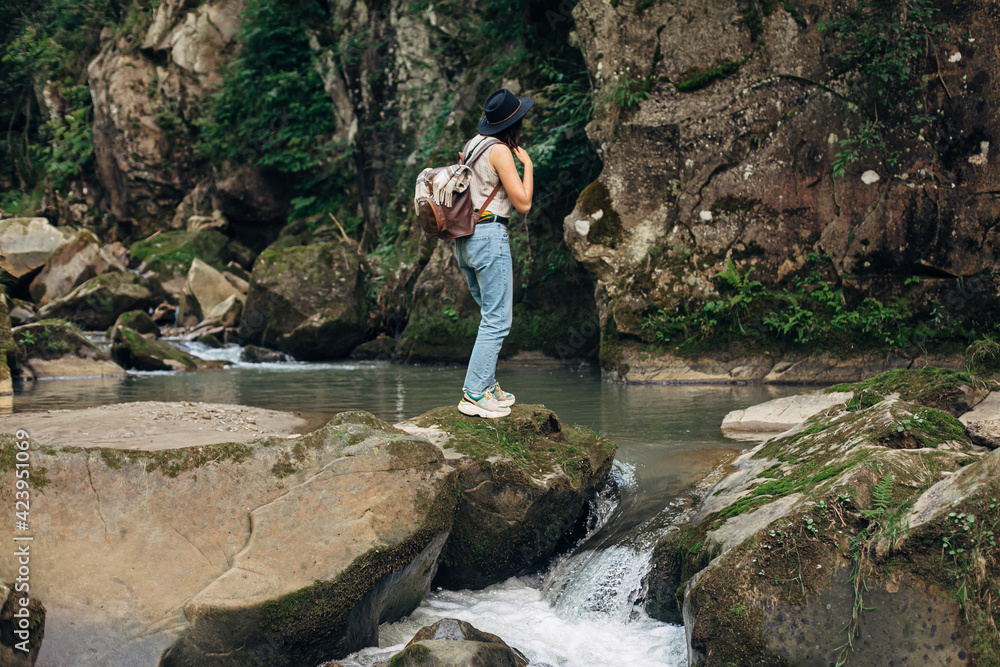 Stylish woman traveler with backpack relaxing at river in mountains. Travel and wanderlust