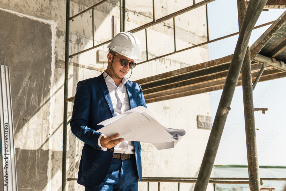 Man architect wearing formal suit and hard hat during building construction control holding a blueprints on a construction site Man architect wearing formal suit and hard hat during building construction control holding a blueprints on a construction site