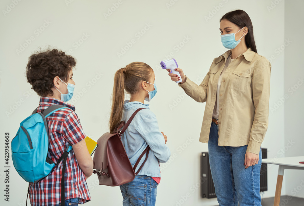 Young female teacher wearing protective mask measuring temperature, screening kids with digital thermometer while students coming to school Young female teacher wearing protective mask measuring temperature, screening kids with digital thermometer while students coming to school