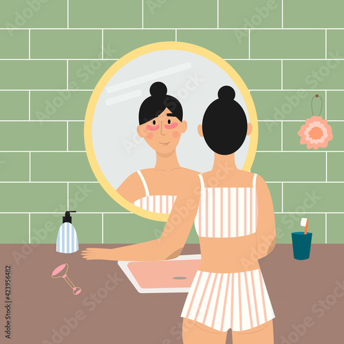 Cute girl in pajamas in the bathroom with reflection in the mirror, with patches under her eyes. Facial massager, body sponge, soap bottle. Morning and evening routines.