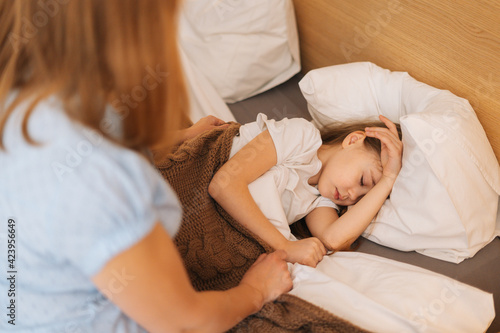 Adorable sick little girl sleeping on bed in children room under blanket at home. Loving young mother sits beside her daughter, worrying but hopeful. Concept of children health.