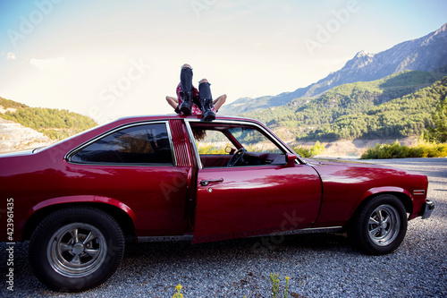 Young carefree woman wearing black ripped jeans  flannel shirt and moto boots lying on the roof of old cherry red muscle car enjoying the mountain view. Traveling alone concept. Copy space  background