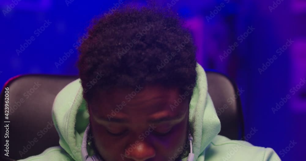 Close up of happy African American young handsome guy dj sitting in dark room recording home studio in neon light, looking at camera in good mood and winking. Male dj composing music in house Close up of happy African American young handsome guy dj sitting in dark room recording home studio in neon light, looking at camera in good mood and winking. Male dj composing music in house