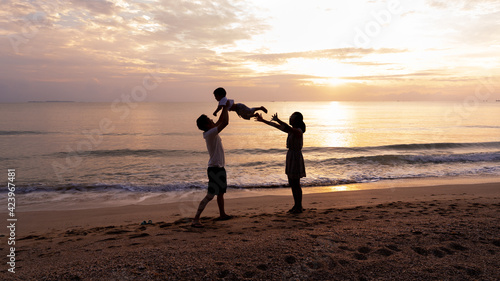 Silhouette picture of father and mother playing with toddler son with happiness moment on the beach in the background of sunrise in the morning time, concept of vacation time of family lifestyle.