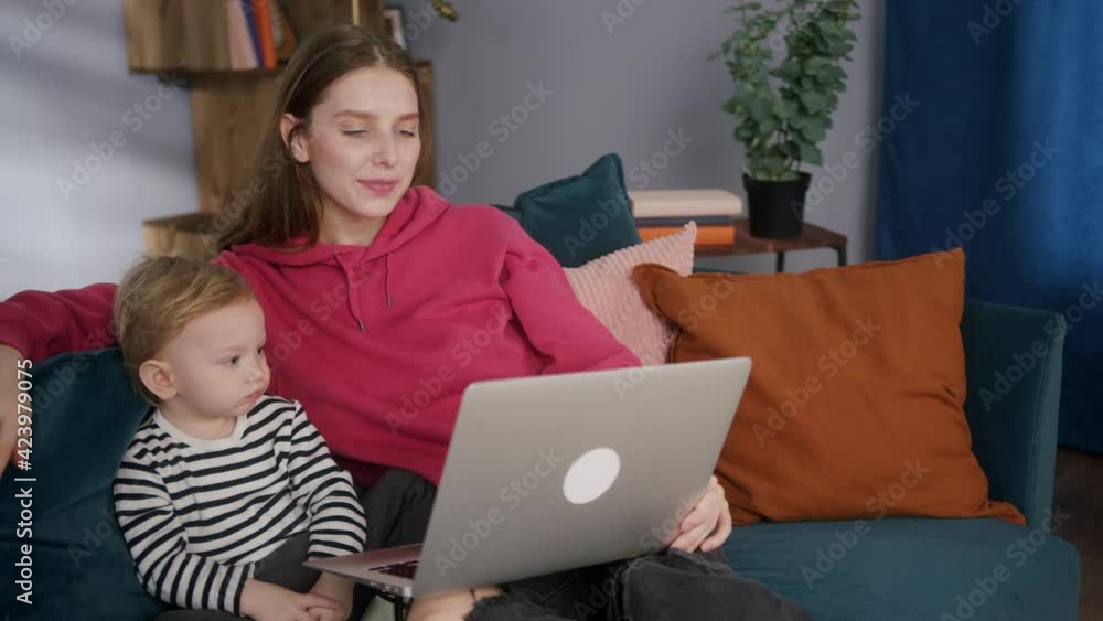 Happy young mother and little son are communicating with friends online, being lockdown. Mom and child are sitting on sofa, using laptop, speaking on video chat with relatives. Happy young mother and little son are communicating with friends online, being lockdown. Mom and child are sitting on sofa, using laptop, speaking on video chat with relatives.