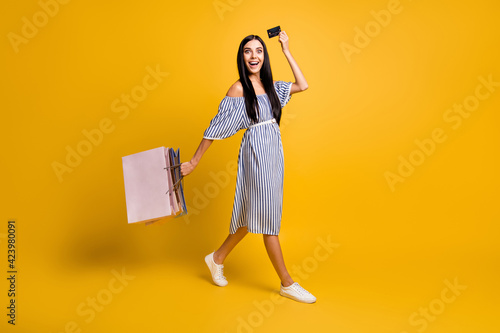 Full length photo of funny excited young lady wear striped dress holding bargains bank card walking isolated yellow color background