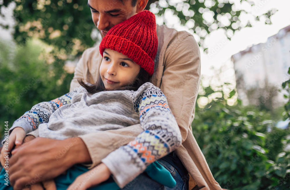 Obraz premium Loving dad holding in his arms his daughter, sitting outdoors. Father embracing his child enjoying the time together. Daddy and daughter share love. Father's day.
