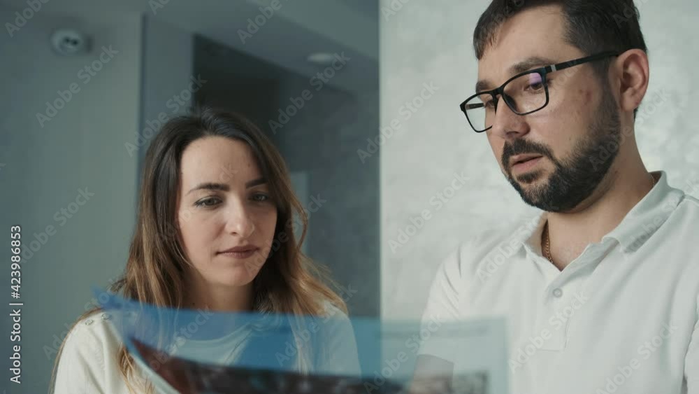 30-year-old dentist man standing talking to brunette patient on sofa and showing tooth x-ray 30-year-old dentist man standing talking to brunette patient on sofa and showing tooth x-ray