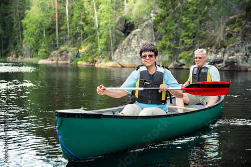 Happy mature couple in life vests canoeing in forest lake. Sunny summer day. Tourists traveling in Finland, having adventure. 