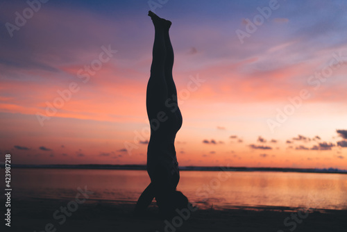 Strong female with athletic body standing on hands and head in asana stretching muscles, woman practice yoga meditation at seashore during evening keeping healthy lifestyle for physical wellness