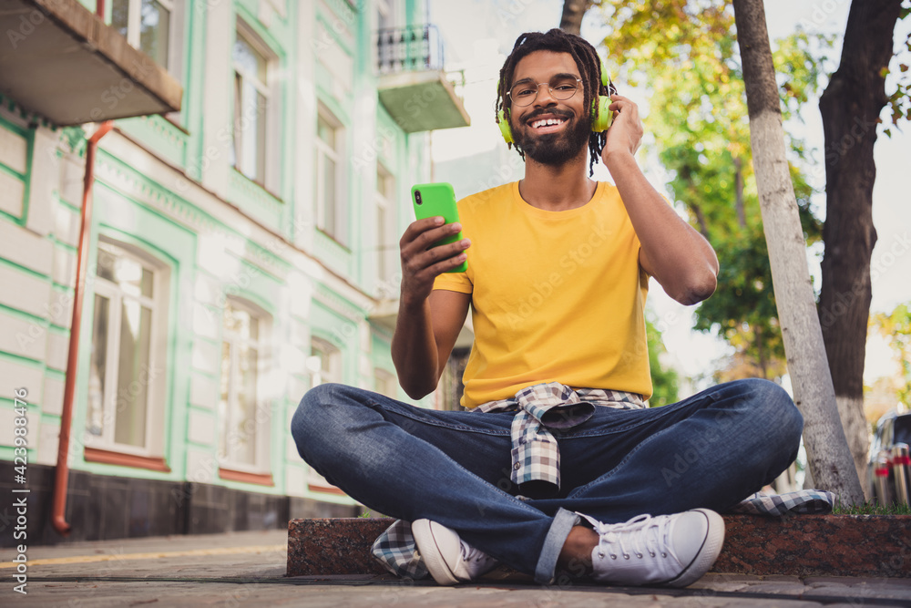 Photo portrait of guy sitting on the ground using smartphone in spectacles drinking beverage listening music headphones - obrazy, fototapety, plakaty 