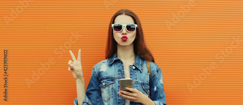 Portrait close up of young woman with smartphone wearing a denim jacket posing on an orange background