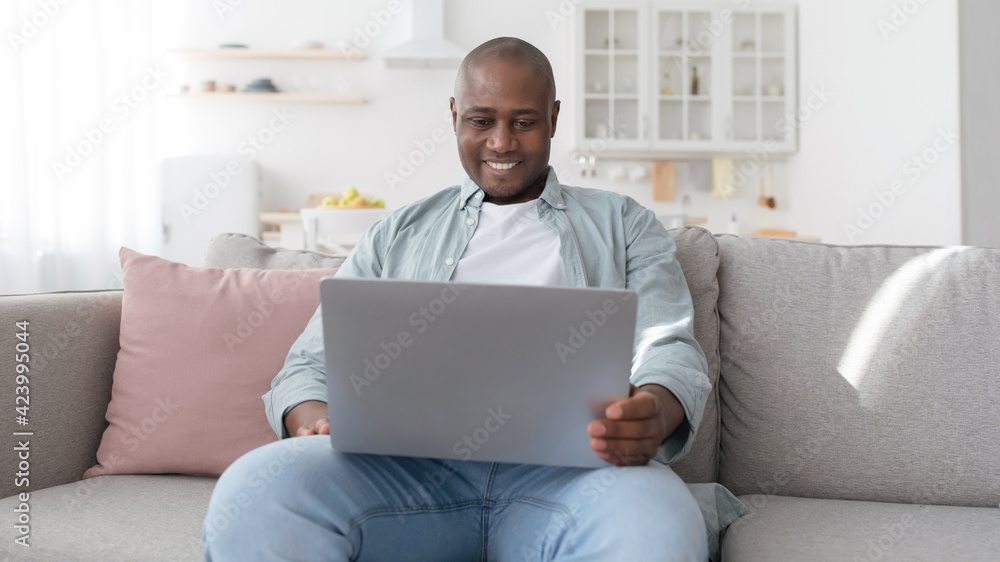 Happy middle-aged black man using laptop while resting on sofa in living room, panorama Happy middle-aged black man using laptop while resting on sofa in living room, panorama