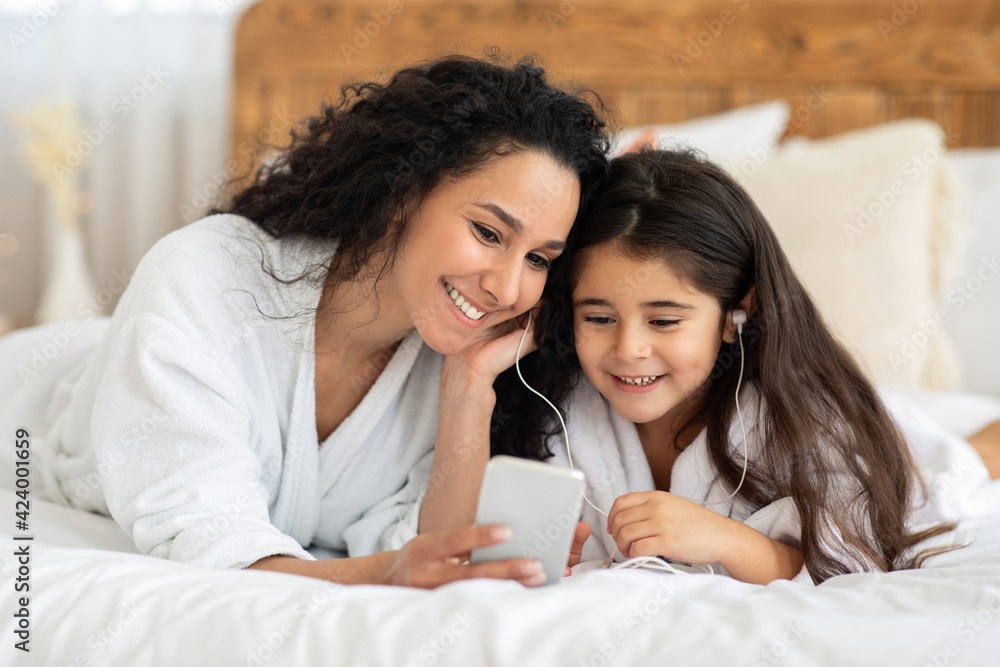 Smiling mom and little girl laying on bed, using smartphone Smiling mom and little girl laying on bed, using smartphone