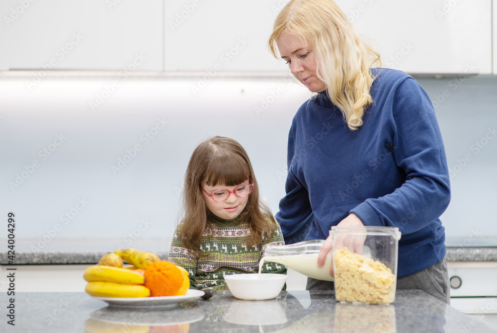 Mom serves breakfast cereal with milk to the girl with down syndrome at home in the kitchen. Ordinary childcare in a family for children with disabilities