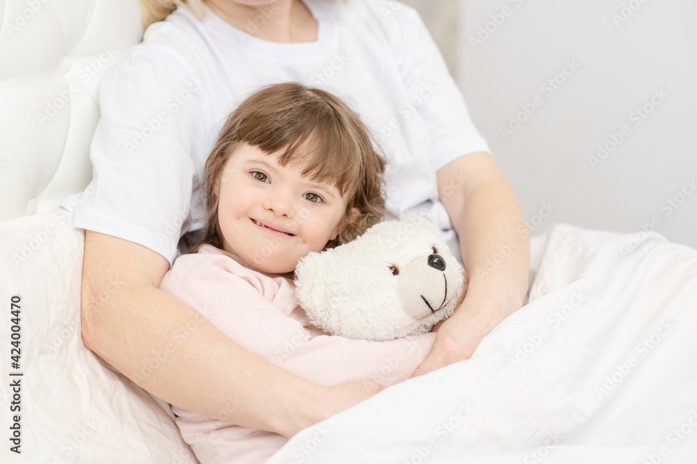 Mom hugging a girl with down syndrome holding a teddy bear at home in the bedroom on the bed. Ordinary childcare in a family for children with disabilities Mom hugging a girl with down syndrome holding a teddy bear at home in the bedroom on the bed. Ordinary childcare in a family for children with disabilities